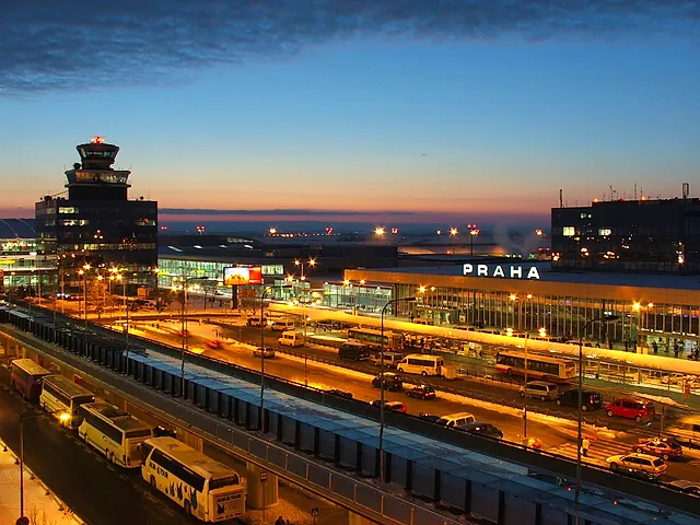 Der Flughafen Prag Václav Havel in der Dämmerung: der Kontrollturm und das Terminal mit dem beleuchteten Schriftzug PRAHA, Busse und Autos auf dem Vorplatz unter einem düsteren Winterhimmel.
