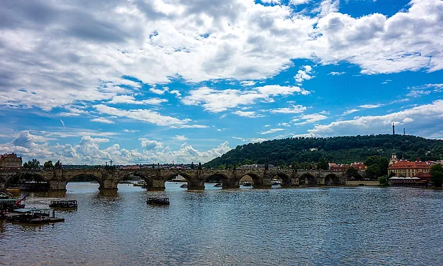 Die steinernen Bögen der Karlsbrücke über die Moldau unter bewölktem blauem Himmel, mit dem bewaldeten Petřín und seinem Aussichtsturm, die hinter der Brücke aufragen.