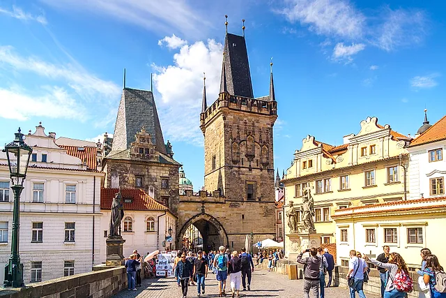 Der gotische Brückenturm der Karlsbrücke mit hohem spitzem Dach und einem Bogendurchgang darunter, Touristen auf der Kopfsteinbrücke und Barockstatuen neben dem Turm.