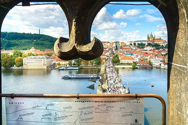 Blick aus dem Inneren eines steinernen Brückenturms durch einen dunklen Eisenbogen auf die Moldau, die Karlsbrücke und rotbedachte Häuser, mit Burgtürmen rechts.