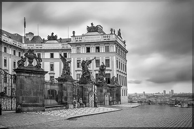 Schwarz-weiße Ansicht eines Barockpalastes hinter schmiedeeisernen Toren und steinernen Statuengruppen, mit gestreiften Wachhäuschen und grauem Himmel über der fernen Stadt.