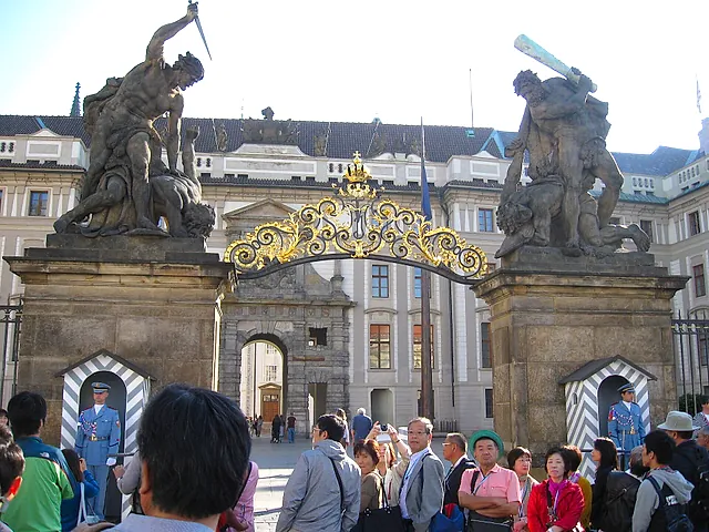 Ein reich verziertes vergoldetes schmiedeeisernes Tor unter einem Barockbogen, flankiert von zwei steinernen Statuen kämpfender Giganten, davor Touristen auf dem Kopfsteinpflaster.