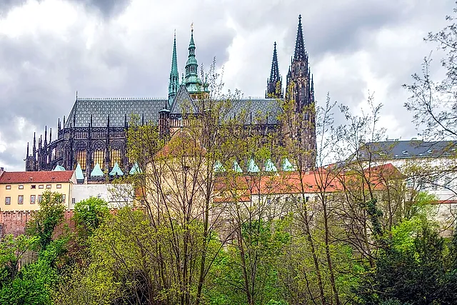 Weite Ansicht der gotischen Türme und Dächer des Veitsdoms, die links über Bäume und blasse Häuser aufragen, unter dramatisch bewölktem Himmel.
