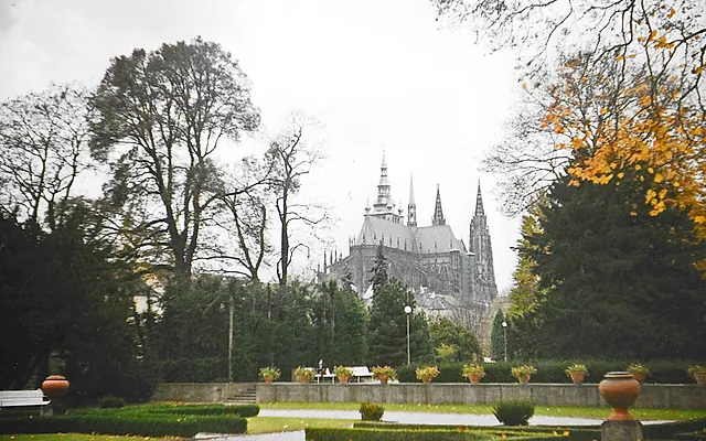 Verblasstes Farbfoto des Veitsdoms über dunklen Bäumen, mit einer gestutzten Hecke, Steinurnen und einer niedrigen Gartenmauer im Vordergrund.