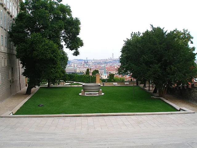 Rasen in einem steingefassten Garten mit einem runden steinernen Brunnenbecken in der Mitte, hohen Bäumen auf beiden Seiten und Blick auf die Stadtdächer dahinter.