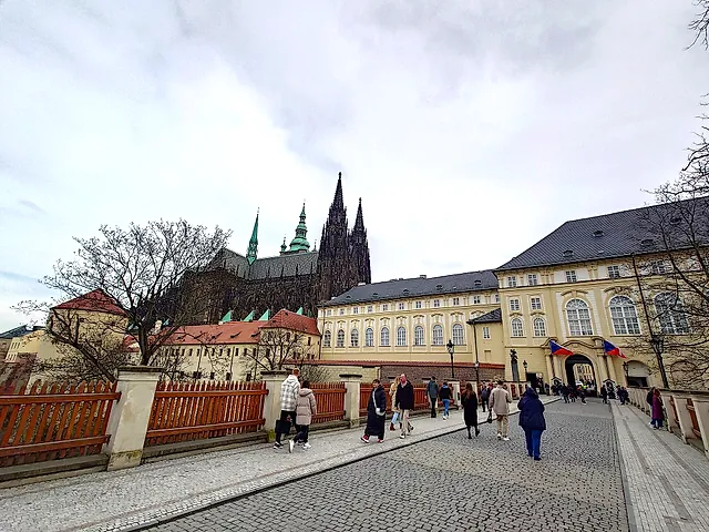 Gepflasterte Straße neben einem Holzzaun, mit den Domtürmen hinter einem gelben Barockpalast mit tschechischen Flaggen und Besuchern, die unter bewölktem Himmel vorbeigehen.