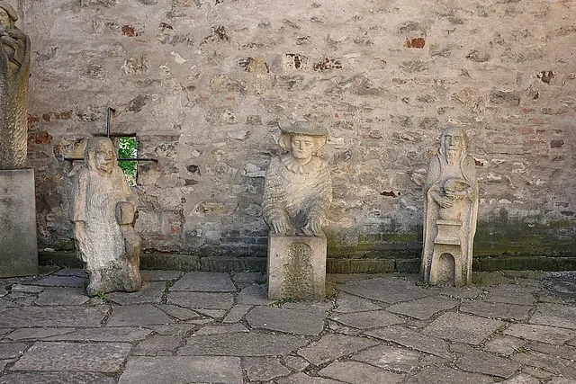 Verwitterte Steinfiguren auf Sockeln vor einer rauen Steinmauer mit einem kleinen Fenster, auf einem unebenen Plattenpflaster in einem Hof.