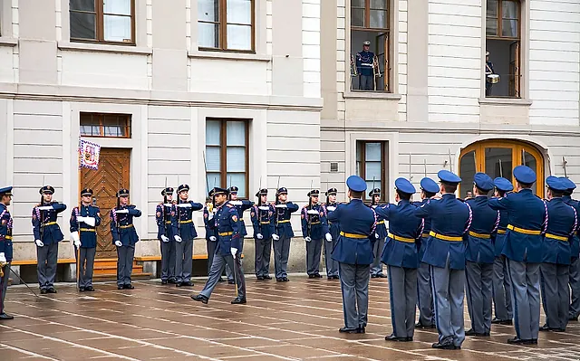 Wachsoldaten in dunkelblauen Paradeuniformen mit weißen Handschuhen stehen in zwei Reihen auf einem nassen Hof vor einem cremefarbenen Palast mit hohen Fenstern und einer gewölbten Holztür.