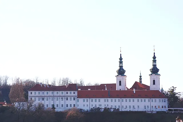 Das Kloster Strahov von unten gesehen, seine lange weiße Barockfassade mit Fensterreihen, roten Dächern und zwei Türmen mit Zwiebelhauben vor blassem Himmel.