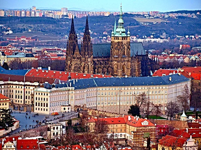 Die Prager Burg vom Petřín aus: die dunklen gotischen Domtürme und die lange Palastreihe erheben sich im weichen Licht über den orangefarbenen Dächern der Kleinseite.