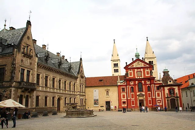 Platz auf der Prager Burg mit der rot-weißen Barockfassade der Georgsbasilika und ihren zwei hellen Türmen, einem reich verzierten Giebelhaus links und Besuchern auf dem Kopfsteinpflaster.