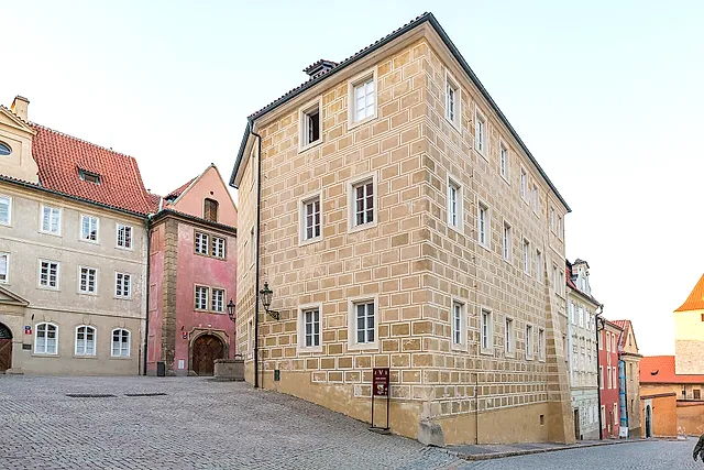 Ecke eines cremefarbenen Hauses mit Rustika-Mauerwerk und Reihen kleiner Fenster, daneben ein rosa Gebäude, an einer Kopfsteingasse unter blassem Himmel.