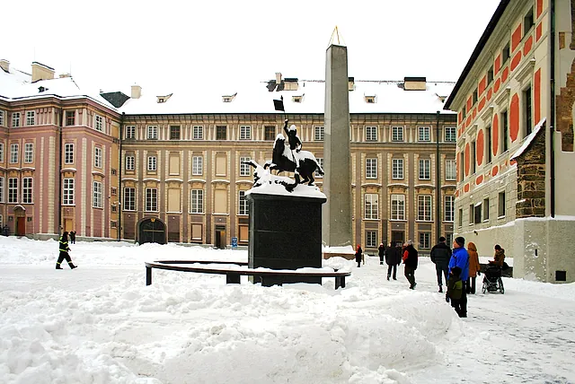 Verschneiter Hof mit einer schwarzen Reiterstatue des heiligen Georg auf einem Steinsockel, einem hohen grauen Obelisken dahinter und Barockfassaden rund um den Platz.