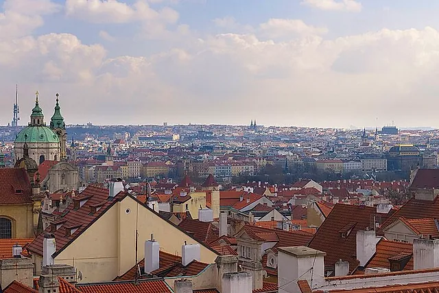 Panoramablick von der Burg über rote Ziegeldächer und eine grüne Kupferkuppel, mit der Stadt, die sich bis zu modernen Türmen am Horizont unter blassem Himmel erstreckt.