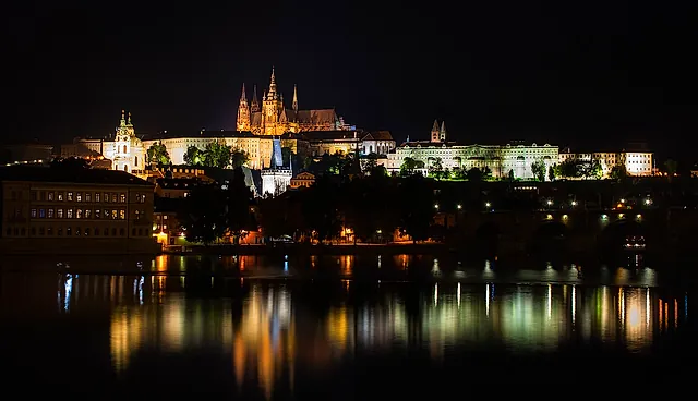 Die Prager Burg und das Ufer bei Nacht: Die Domtürme und Palastfassaden sind über dunklen Dächern angestrahlt, und das Wasser spiegelt die Lichter.