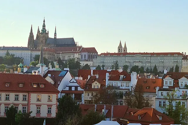 Prague Castle and the spires of St Vitus Cathedral at sunset, seen across the Lesser Town rooftops, with a long line of palace facades along the hilltop.