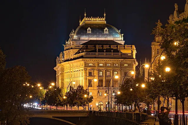 Das Nationaltheater in Prag bei Nacht, seine Neorenaissance-Fassade in warmem Licht, darüber ein Kuppeldach, mit Bäumen und Laternen am Ufer.