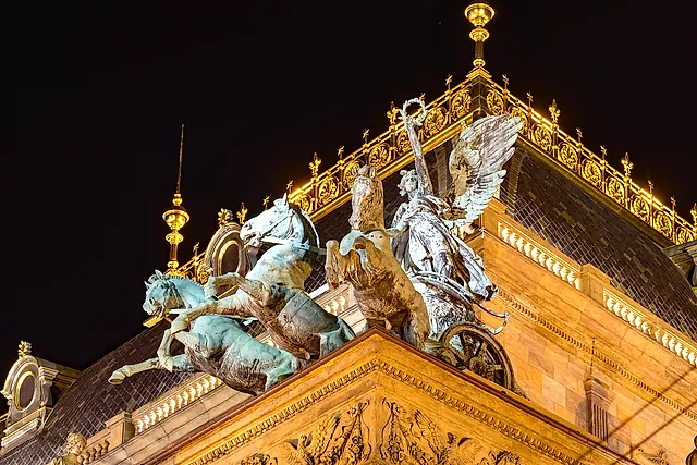 Vergoldeter Schmuck und eine bronzene Quadriga auf dem Dach des Nationaltheaters bei Nacht, von unten angestrahlt vor dunklem Himmel.