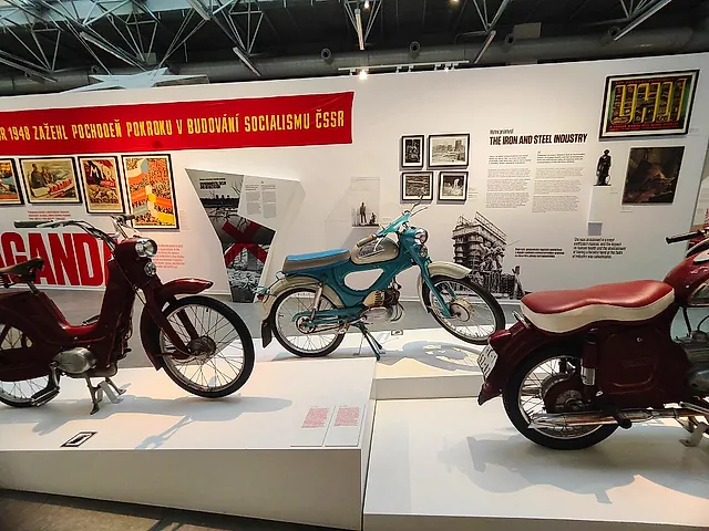 Exhibition room of the Museum of Communism in Prague, with a red banner in Czech lettering, vintage motorbikes on white plinths and display panels on the wall.