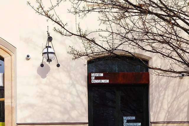 The entrance of the Museum of Communism in Prague: a rust-coloured sign reading MUSEUM OF COMMUNISM above a dark glass door under an arch, on a cream wall with a globe lamp and the shadows of bare branches.