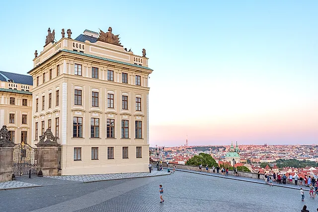 Die Ecke eines gelben Barockpalastes auf der Prager Burg mit geschwungener Dachlinie, daneben eine gepflasterte Terrasse, auf der Besucher am Geländer entlanggehen; dahinter die Stadt und eine Kirchenkuppel.