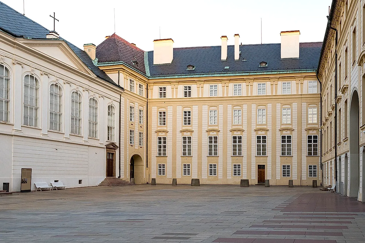 Il Secondo Cortile del Castello di Praga, un ampio piazzale lastricato bordato da facciate barocche color crema, con finestre alte e alcune panchine bianche, sotto un cielo chiaro.