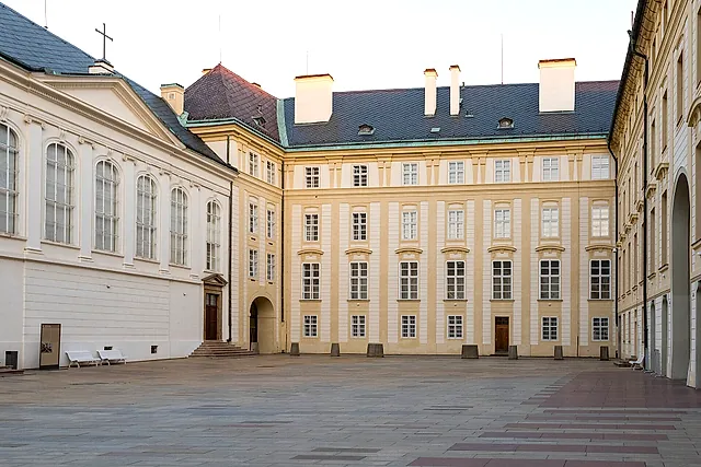 The Second Courtyard of Prague Castle, a wide paved square lined with cream Baroque facades, tall windows and a few white benches, under a pale sky.