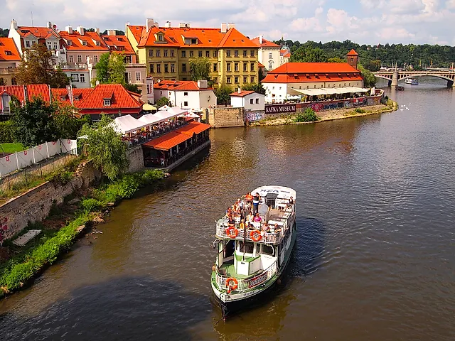 Ein weißes Ausflugsboot mit grünen Akzenten und Passagieren auf dem offenen Deck fährt auf der braunen Moldau, mit rotbedachten Häusern und einer Brücke dahinter.