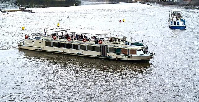 Ein langes weißes Fahrgastschiff, sein Name am Rumpf, quert die Moldau unter grauem Himmel, mit Passagieren auf dem Oberdeck und einem kleinen blauen Boot dahinter.