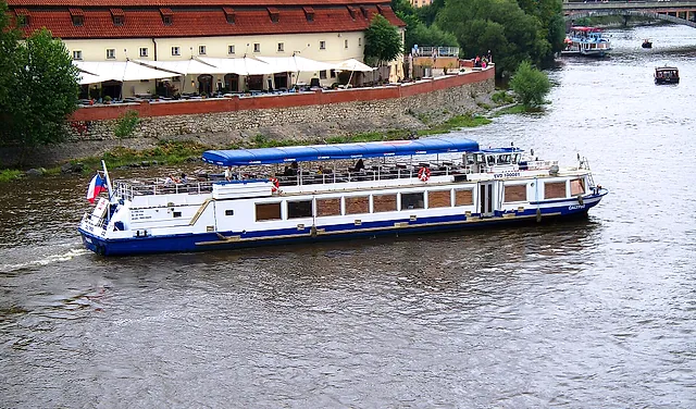 Ein weißes zweistöckiges Ausflugsschiff mit blauem Sonnensegel fährt auf der Moldau an einer cremefarbenen Uferterrasse mit weißen Sonnenschirmen vorbei, in der Ferne ein weiteres Boot.
