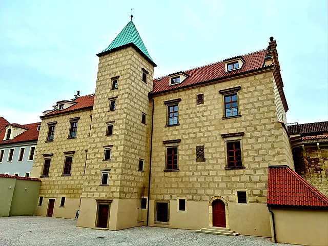 Ein Burggebäude mit quadratischem Eckturm unter grüner Kupferhaube, Mauern mit Quadermuster und rotem Ziegeldach über einem leeren gepflasterten Hof unter hellblauem Himmel.