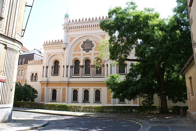 Fassade der Spanischen Synagoge in Prag, ein cremefarbenes Gebäude mit Bogenfenstern und einer Zinnenlinie, teilweise von einem großen Baum in einer ruhigen Straße verdeckt.