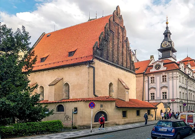 Die Altneuschul in Prag, eine gotische Halle mit Stufengiebel und steilem rotem Ziegeldach, neben einem rosa Gebäude mit Uhrturm unter bewölktem Himmel.