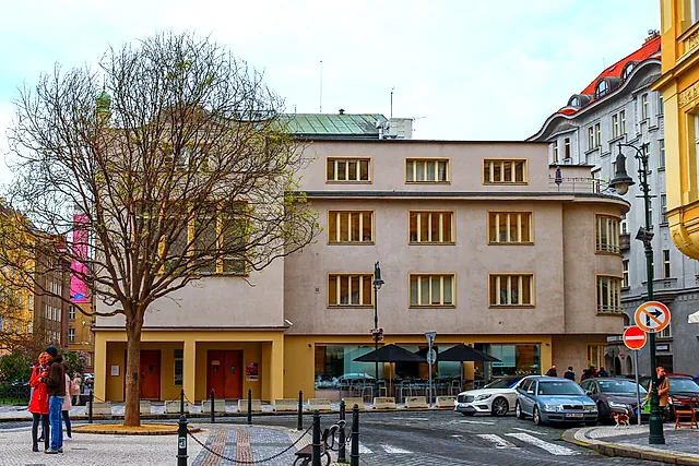 Ein helles Gebäude mit Reihen hoher Fenster an einem Stadtplatz, davor ein kahler Baum, einige Menschen und parkende Autos in der Nähe unter blassem Himmel.