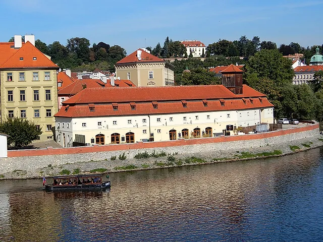 Ein langes cremefarbenes Museumsgebäude mit rotem Ziegeldach und Bogenfenstern am Moldauufer, mit einem Ausflugsboot auf dem Wasser und Bäumen dahinter.