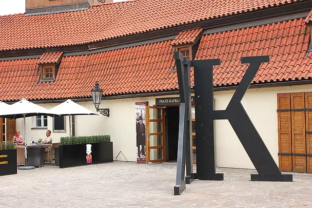 Ein hoher schwarzer stählerner Buchstabe K in einem gepflasterten Hof vor einem cremefarbenen Gebäude mit rotem Ziegeldach, links eine Caféterrasse unter weißen Sonnenschirmen.
