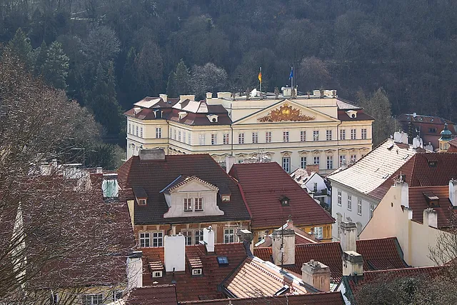 Ein blassgelber Barockpalast mit geschwungenem Mittelgiebel und zwei Flaggen auf dem Dach, über roten Dächern und dunklen bewaldeten Hängen im Winterlicht gesehen.