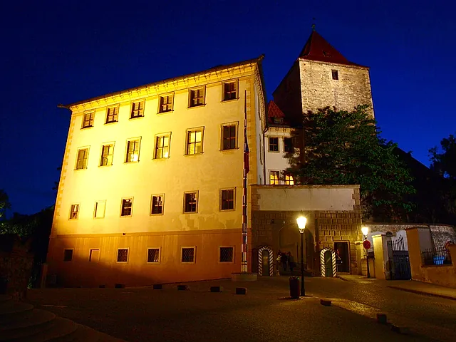 Ein cremefarbener Palast bei Nacht mit Reihen beleuchteter Fenster, einem rotbedachten Steinturm dahinter, einem Fahnenmast und einer Laterne auf dem Kopfsteinplatz.