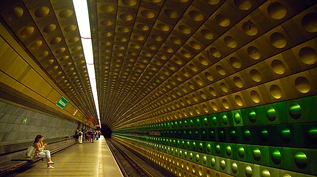 Bahnsteig der Station Malostranská der Prager U-Bahn-Linie A, mit einer geschwungenen Wand aus runden, grün beleuchteten Platten und einigen wartenden Fahrgästen am Bahnsteig.