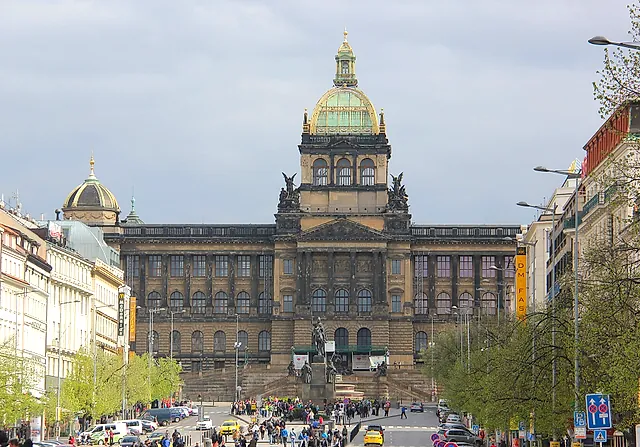 Das Nationalmuseum in Prag, ein großes Gebäude mit grüner Kupferkuppel, unter grauem Himmel gesehen, mit Verkehr auf der Allee und kleinen Gestalten in der Ferne.