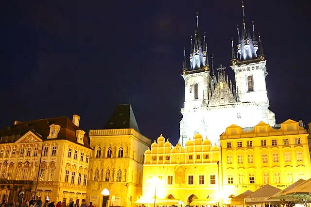Der Altstädter Ring bei Nacht, mit den Zwillingstürmen der Teynkirche über angestrahlten gotischen und barocken Häusern und einigen kleinen Gestalten in den unteren Ecken.