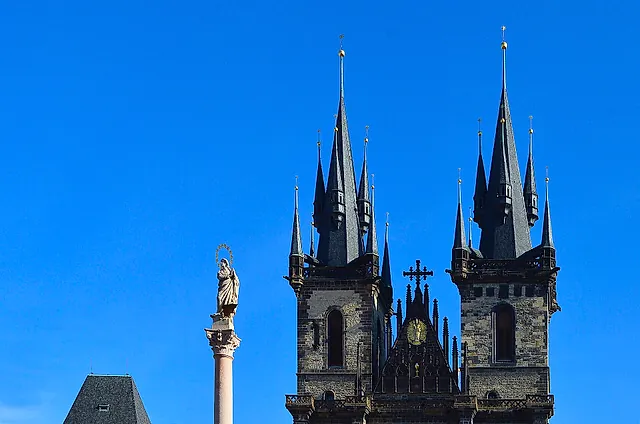 Dunkle gotische Türme der Teynkirche vor tiefblauem Himmel, mit einer Säule mit Statue unten links und Dächern darunter.