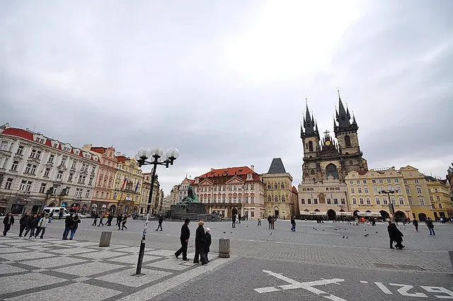 Der Altstädter Ring im grauen Tageslicht, mit den Türmen der Teynkirche rechts und pastellfarbenen Fassaden rund um den Platz und einigen Menschen, die über das Pflaster gehen.