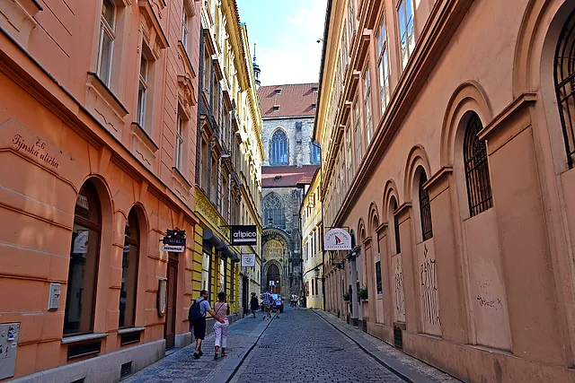 Eine schmale Kopfsteingasse in der Altstadt, mit orangefarbenen und blassgelben Hausfronten, Bogentüren und einer gotischen Kirche am Ende, mit einigen fernen Fußgängern.