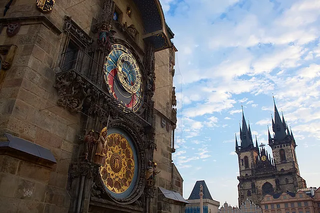 Das astronomische Zifferblatt am Altstädter Rathausturm, dahinter die gotischen Zwillingstürme der Teynkirche vor bewölktem blauem Himmel.