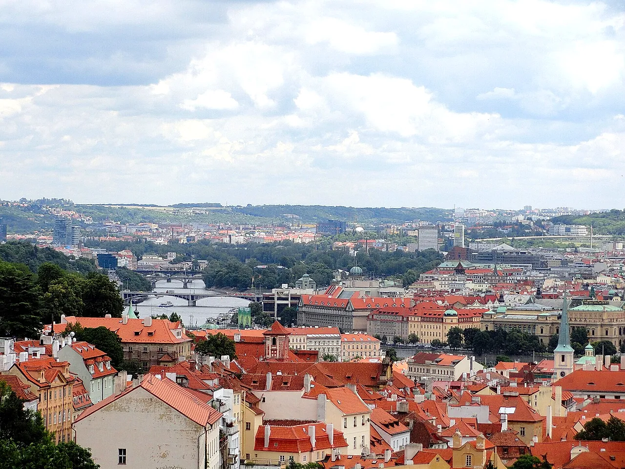 Panoramic view over Prague's red-tiled rooftops toward the Vltava and its bridges, with green hills and modern towers on the far bank under a cloudy sky.
