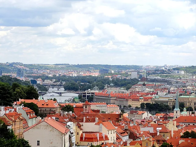 Panoramablick über Prags rote Ziegeldächer zur Moldau und ihren Brücken, mit grünen Hügeln und modernen Türmen am anderen Ufer unter bewölktem Himmel.