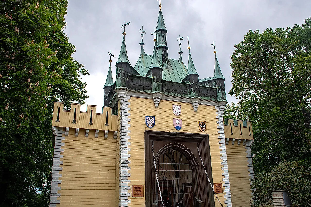 O portão de entrada do Labirinto de Espelhos na colina de Petřín: um edifício amarelo-claro com torreões e telhados de cobre verde, uma porta de madeira e brasões heráldicos acima do arco.