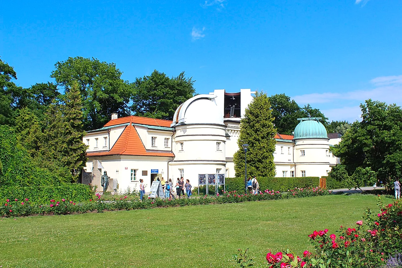 O Observatório Štefánik na colina de Petřín: um edifício branco com cúpula redonda e uma torre de telhado verde, sobre um gramado com rosas vermelhas e alguns visitantes.