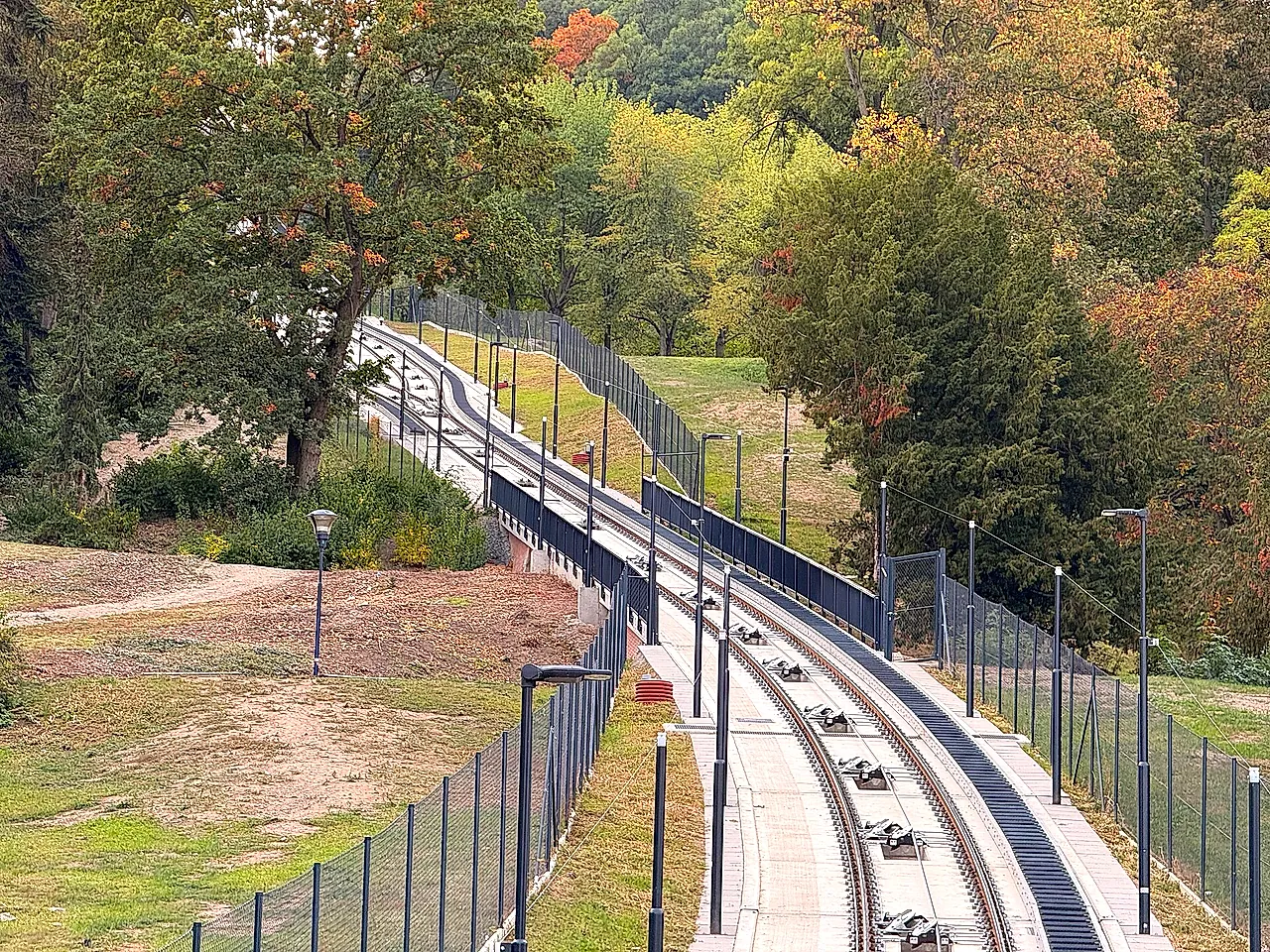 O trilho do funicular subindo uma encosta arborizada da colina de Petřín no outono, com uma cerca de metal ao longo da linha, postes de luz ao lado do caminho e nenhum carro à vista.